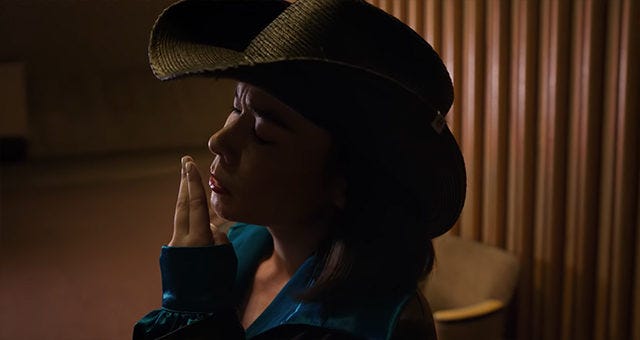 Mitski em Working For The Knife usando chapéu de caubói e fingindo fumar um cigarro