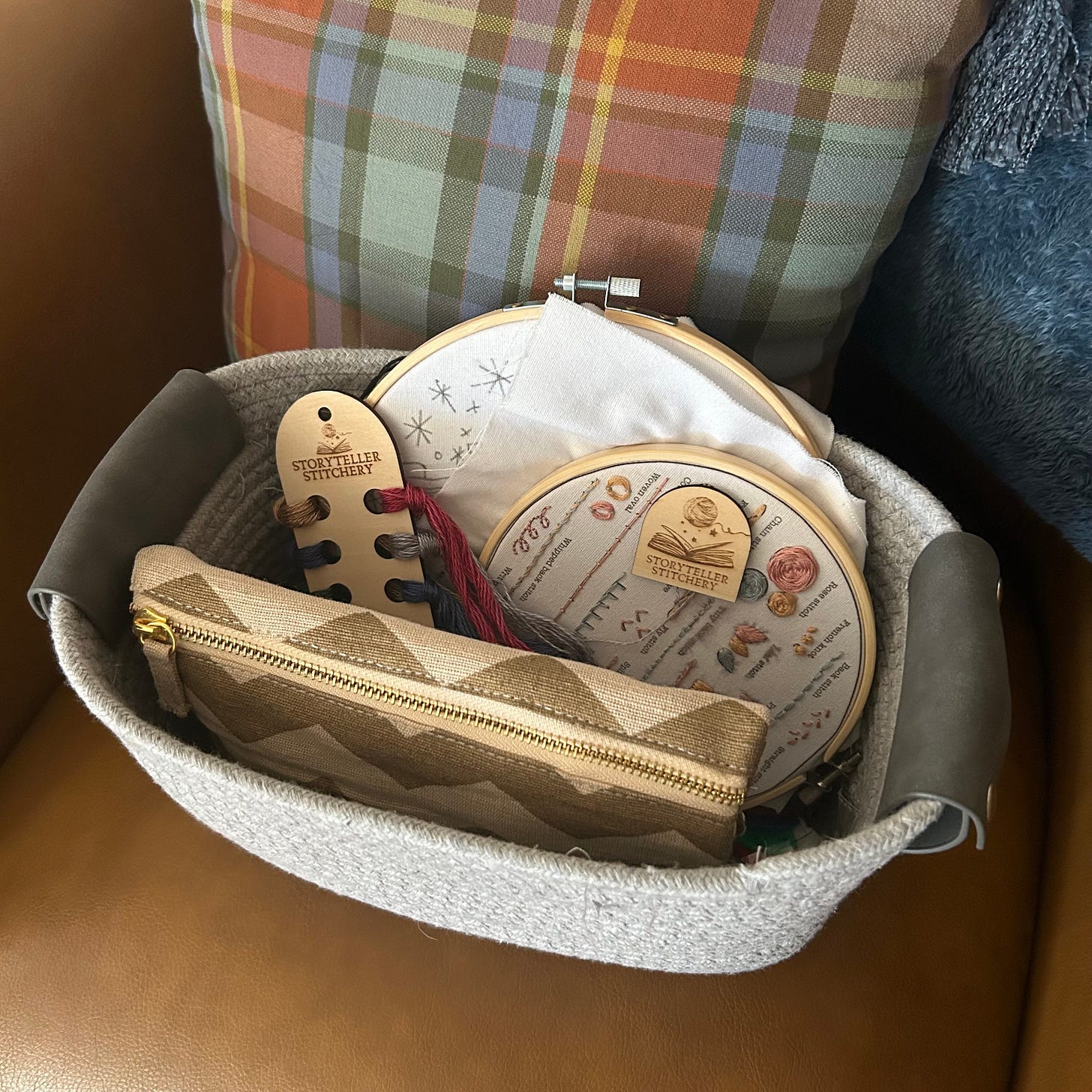 Small grey basket containing embroidery projects and tools, including two hoops with partially stitched patterns, threads, a needle keeper, and a pouch with more tools inside.