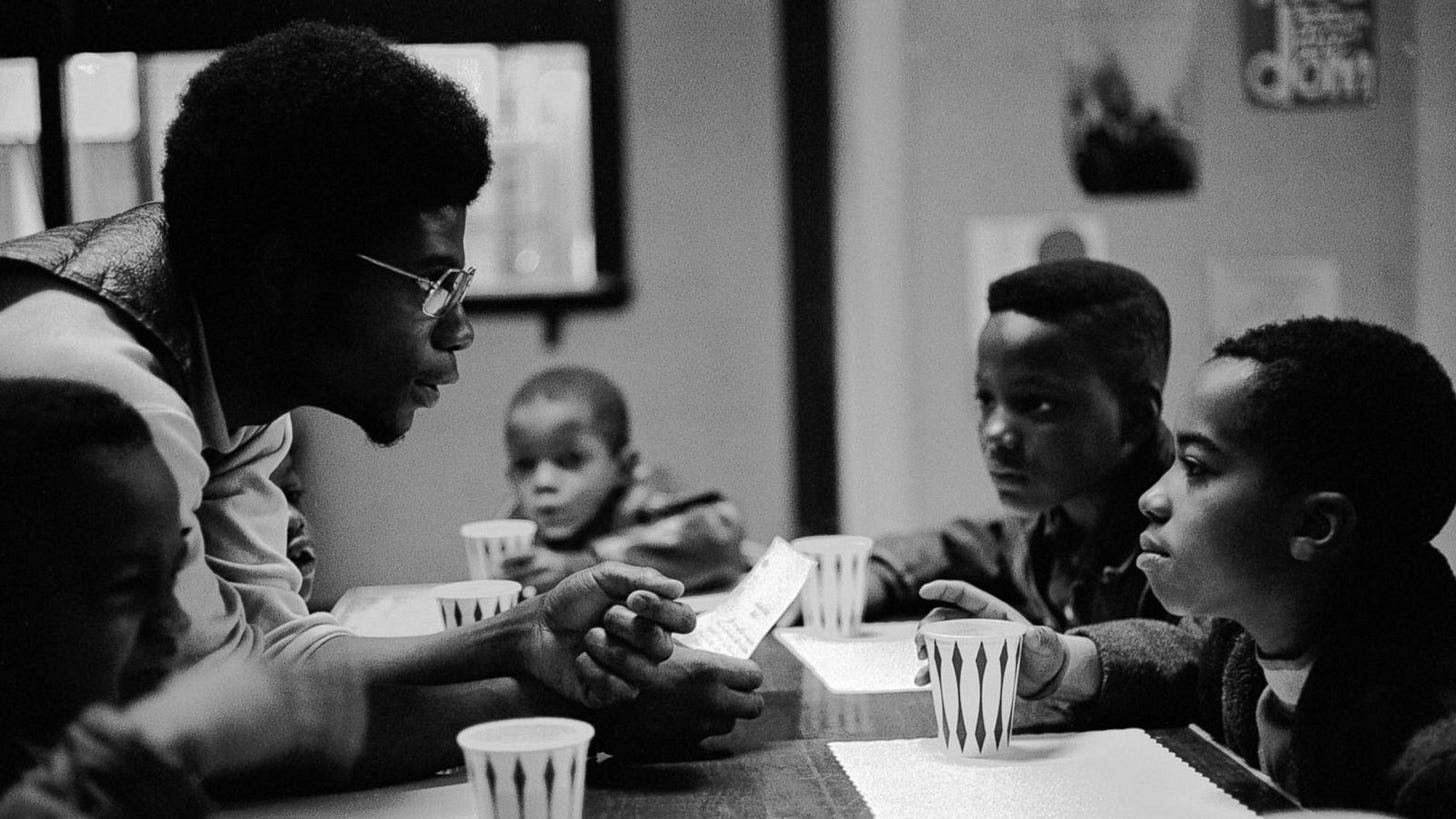 Black and white photo of a Black man wearing glasses talking intensely to two young black boys at a table. It looks they have just finished a meal, because table mats are visible and they each gave cups close by. Some very life lessons are being imparted.
