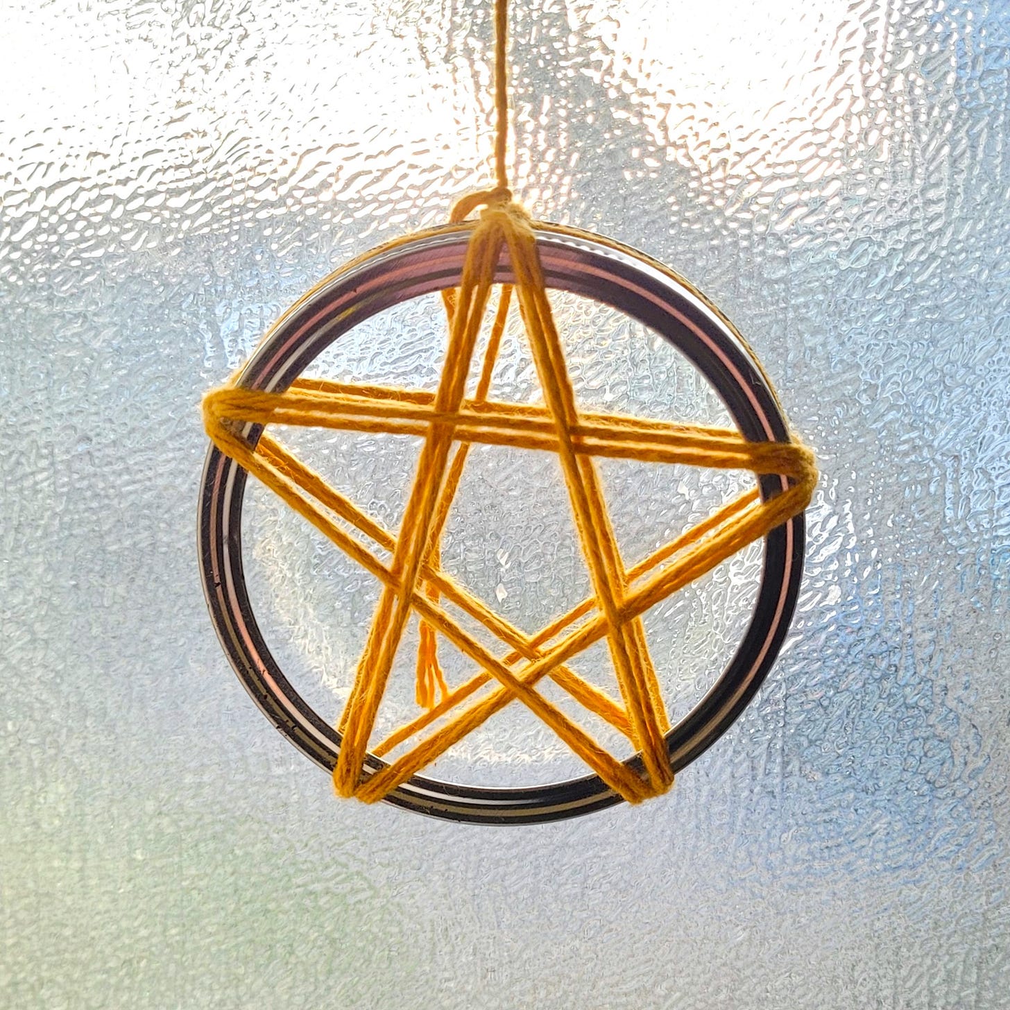 Yellow yarn, wrapped around the metal ring of a mason jar to form a pentagram. It's hanging in front of a sunny window.