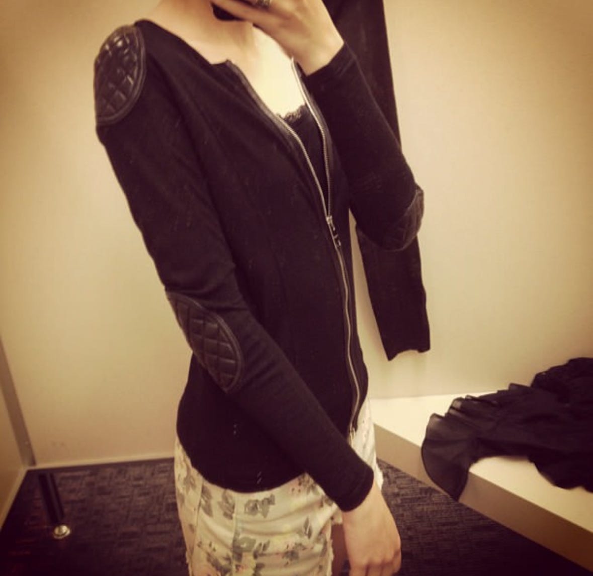 Young woman from neck to knee. Pictured taking a selfie with a Burberry moto style jacket on with elbow patches and some floral shorts.