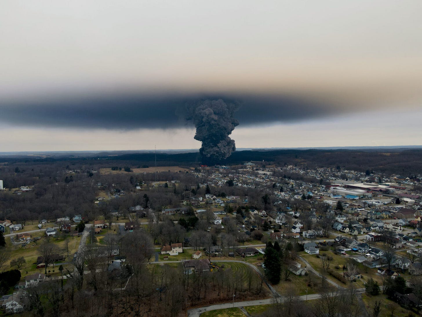 Massive dark smoke plume rising over a East Palestine  under an overcast sky, seen from the air.