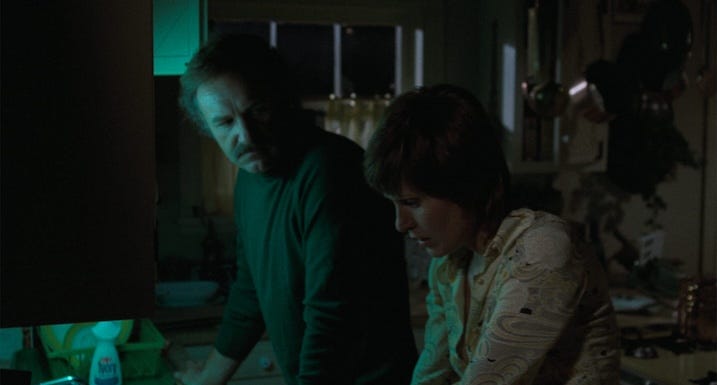 Gene Hackman and Susan Clark stand under dim green lighting in a kitchen, tension thick between them as they speak in low tones.