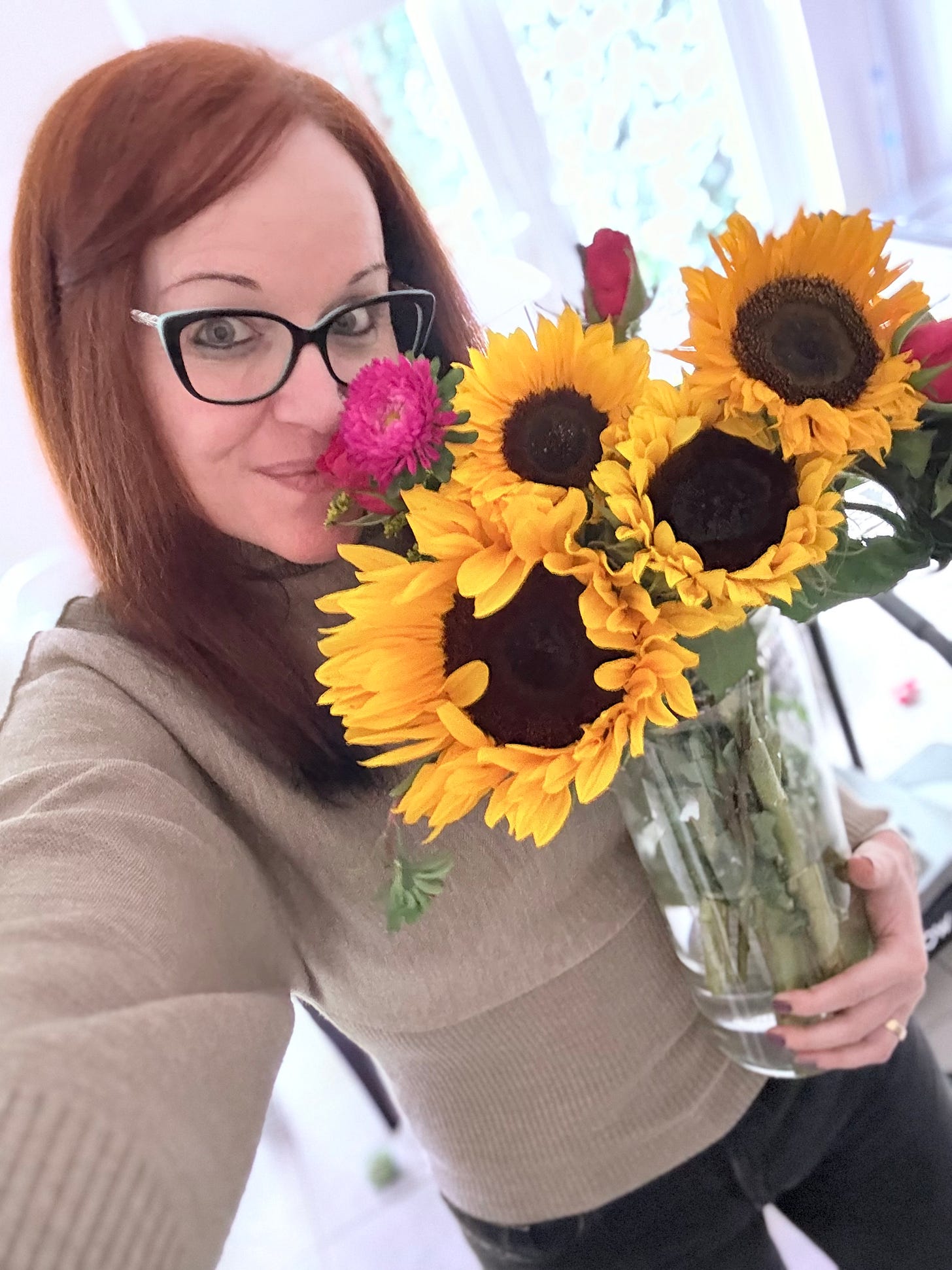 Georgina Dean, wearing a cozy beige sweater, holds a large vase of bright yellow sunflowers, symbolizing the theme of the autumnal shift and harvesting creative ideas.