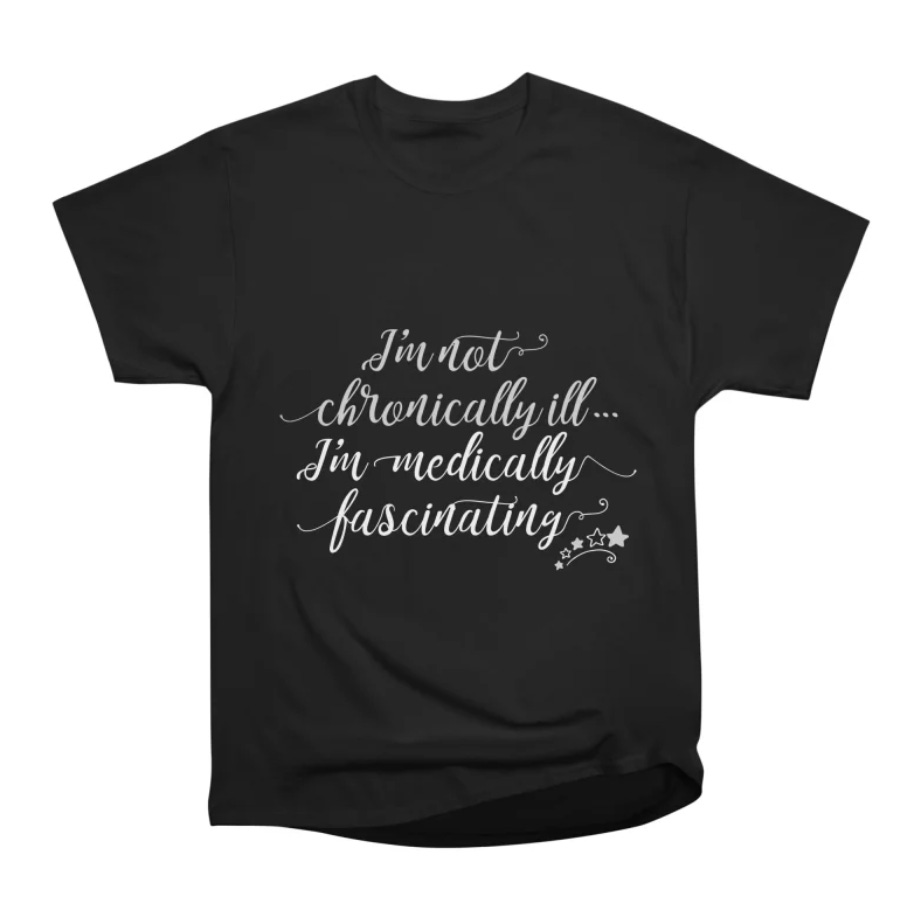 A black short-sleeve T-shirt displayed against a light gray background. Centered on the front is white decorative script that reads, “I’m not chronically ill… I’m medically fascinating,” with small star and sparkle accents beneath the text. The design uses elegant, flowing lettering for a playful, tongue-in-cheek statement about living with chronic illness.