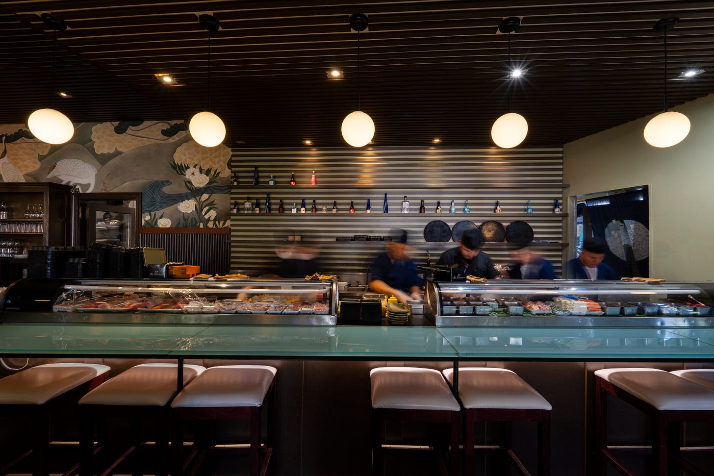 A direct view of a sushi bar, with blurred images of people behind it working, two glass cases side by side on the bar filled with plates of fish, and a wall behind the bar with a shelf across it.