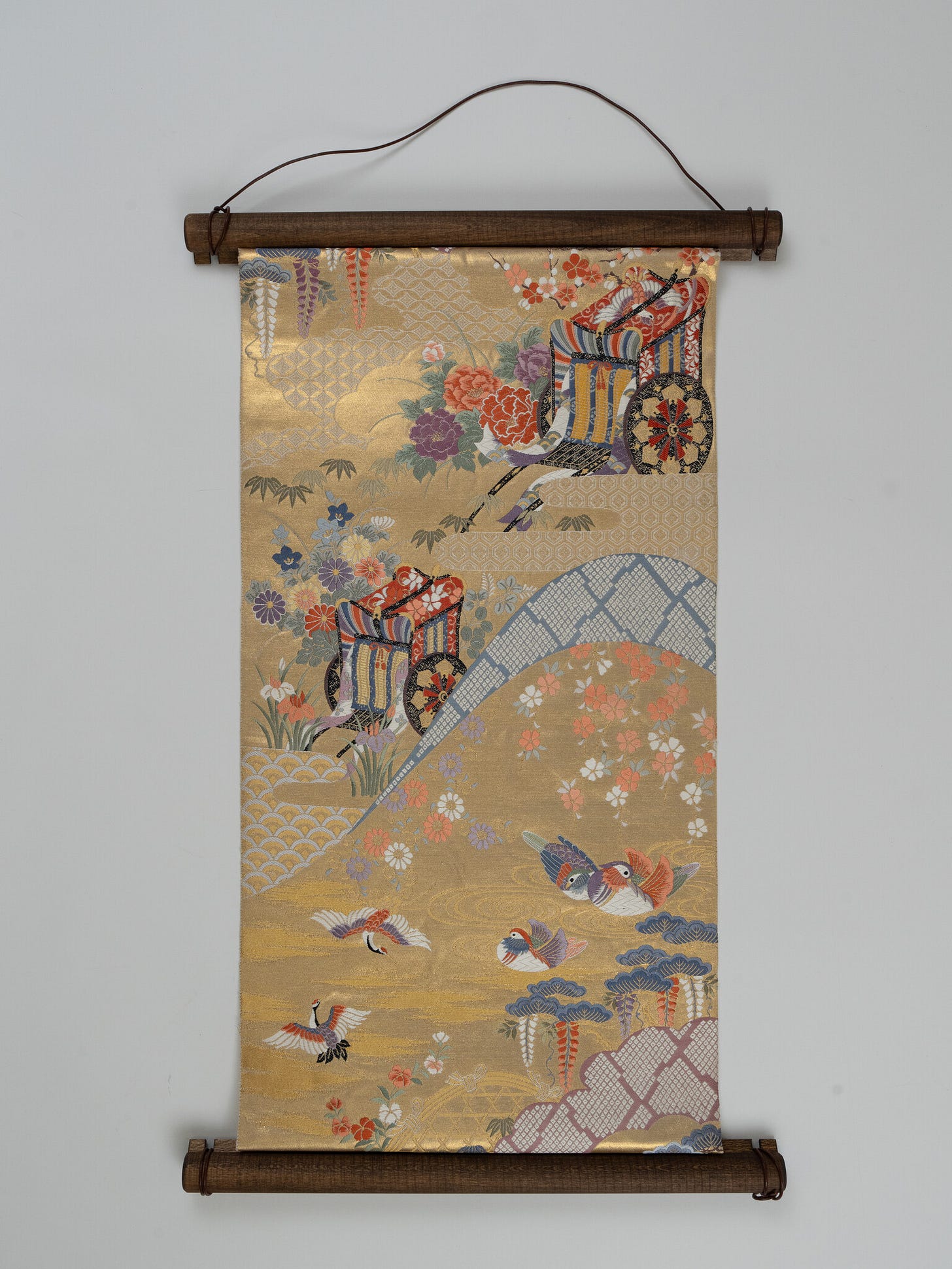 A gold-ground brocade obi hung vertically from a dark wooden rod against a pale grey wall, densely patterned with floral clusters, ox-carts, cranes, paired ducks, and wave-scale motifs. A gold-ground brocade obi hung vertically from a dark wooden rod against a pale grey wall, densely patterned with floral clusters, ox-carts, cranes, paired ducks, and wave-scale motifs.