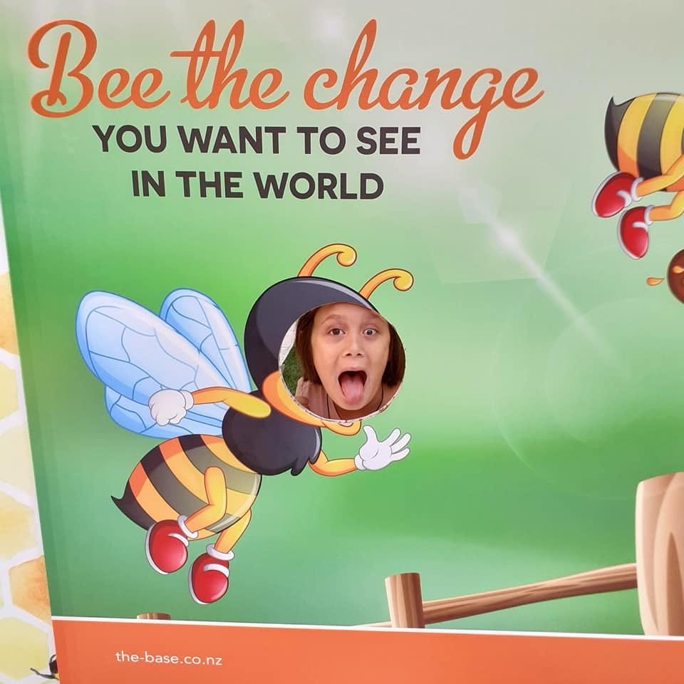 picture of a board that reads, bee the change you want to see in the world. It has an image of a bee, and a cut out where a face can go. A child's face looks out
