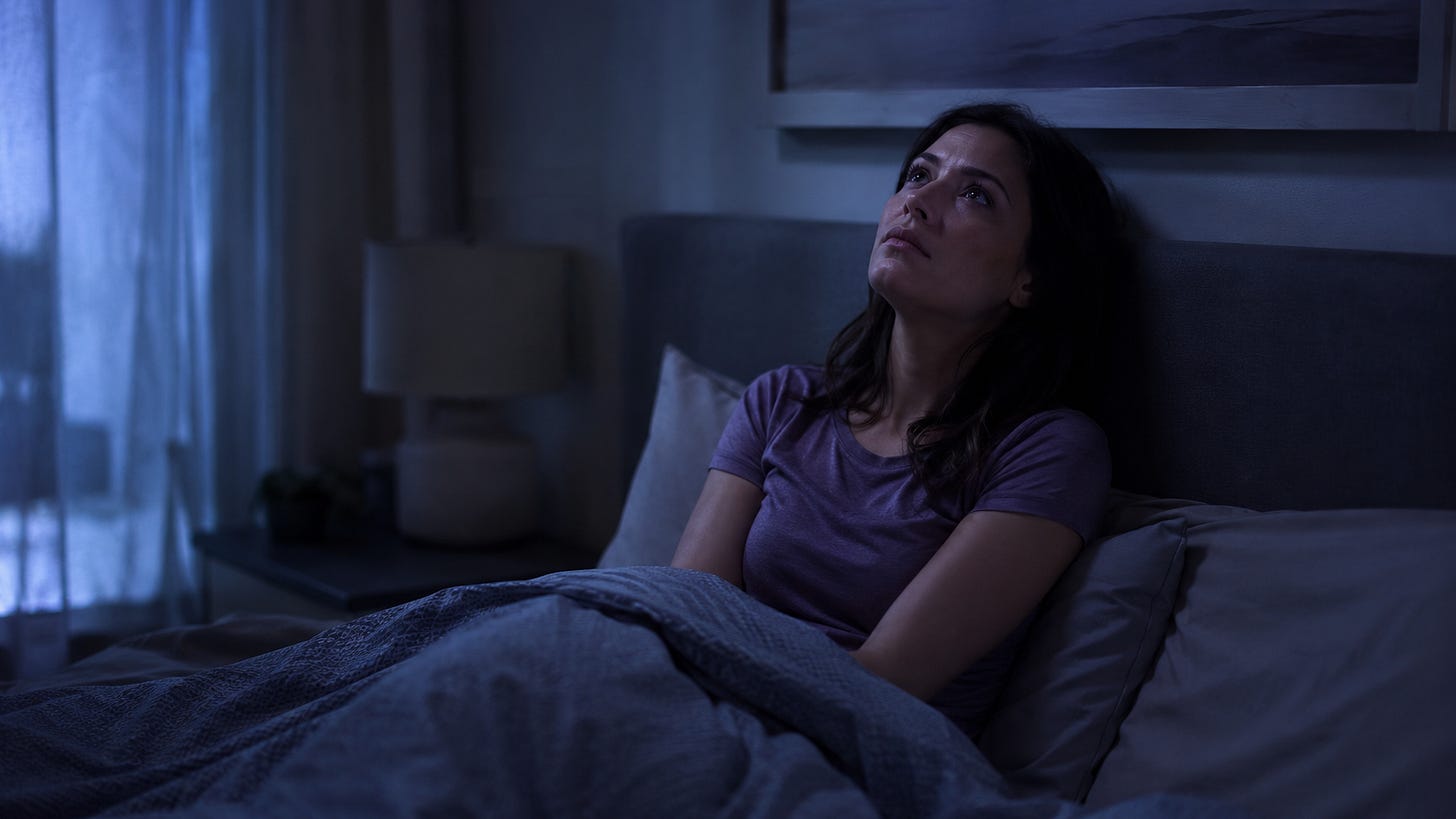 A woman sits upright in bed late at night, staring at the ceiling with a restless expression. The room is dark and quiet, softly lit by faint blue light from a nearby window, creating a calm but sleepless mood.