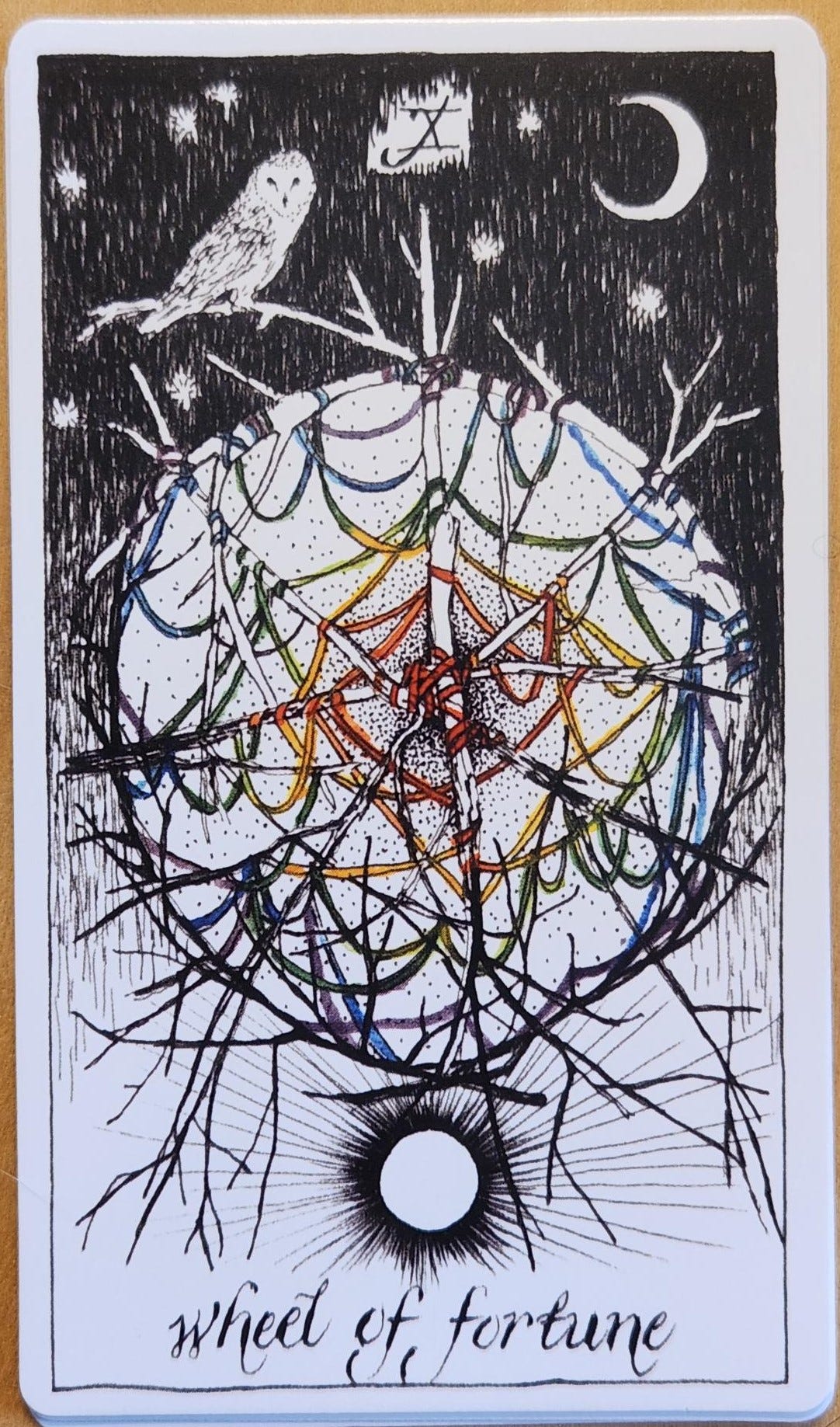 A tarot card labelled X wheel of fortune. The wheel shows branches tied together with colourful threads or green, blue, purple, red and yellow. The threads are criss-crossed and connected across the entire wheel, forming a network. Above the wheel, is a night sky with a crescent moon and an Owl perched upon a branch. Below the wheel, is a day sky with a full sun shining. 