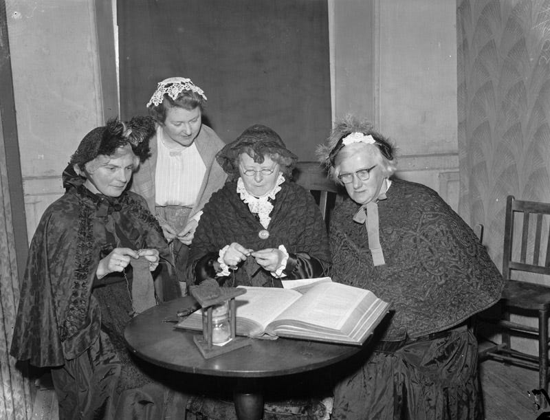 A group of women in traditional Welsh costume gathered for a pageant, sat knitting.