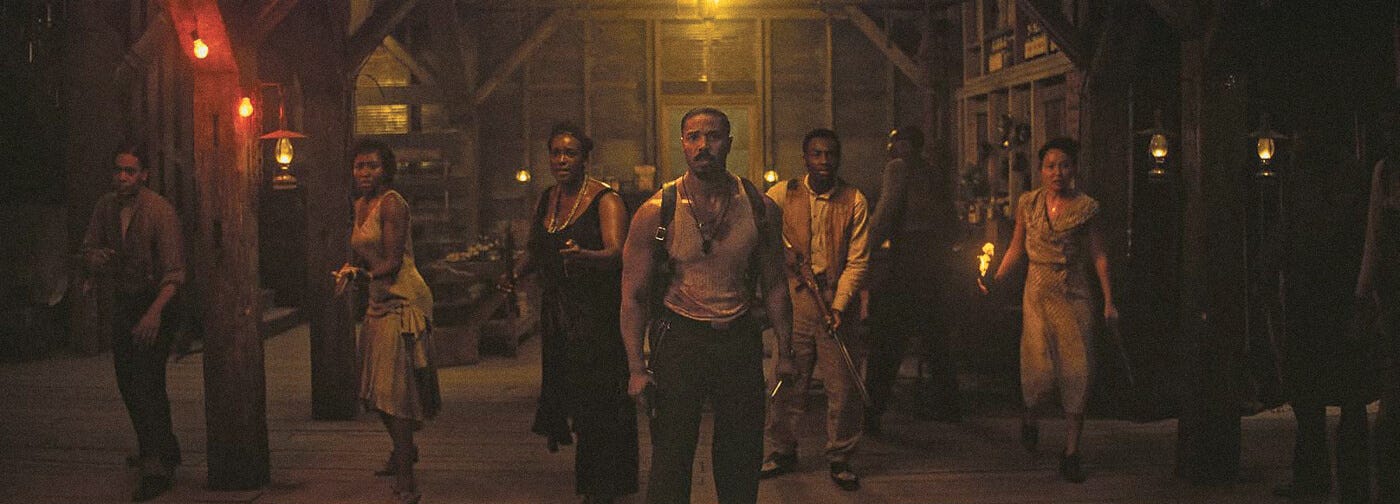 Michael B. Jordan leads a group of townspeople armed with weapons through a lantern-lit barn in Sinners.