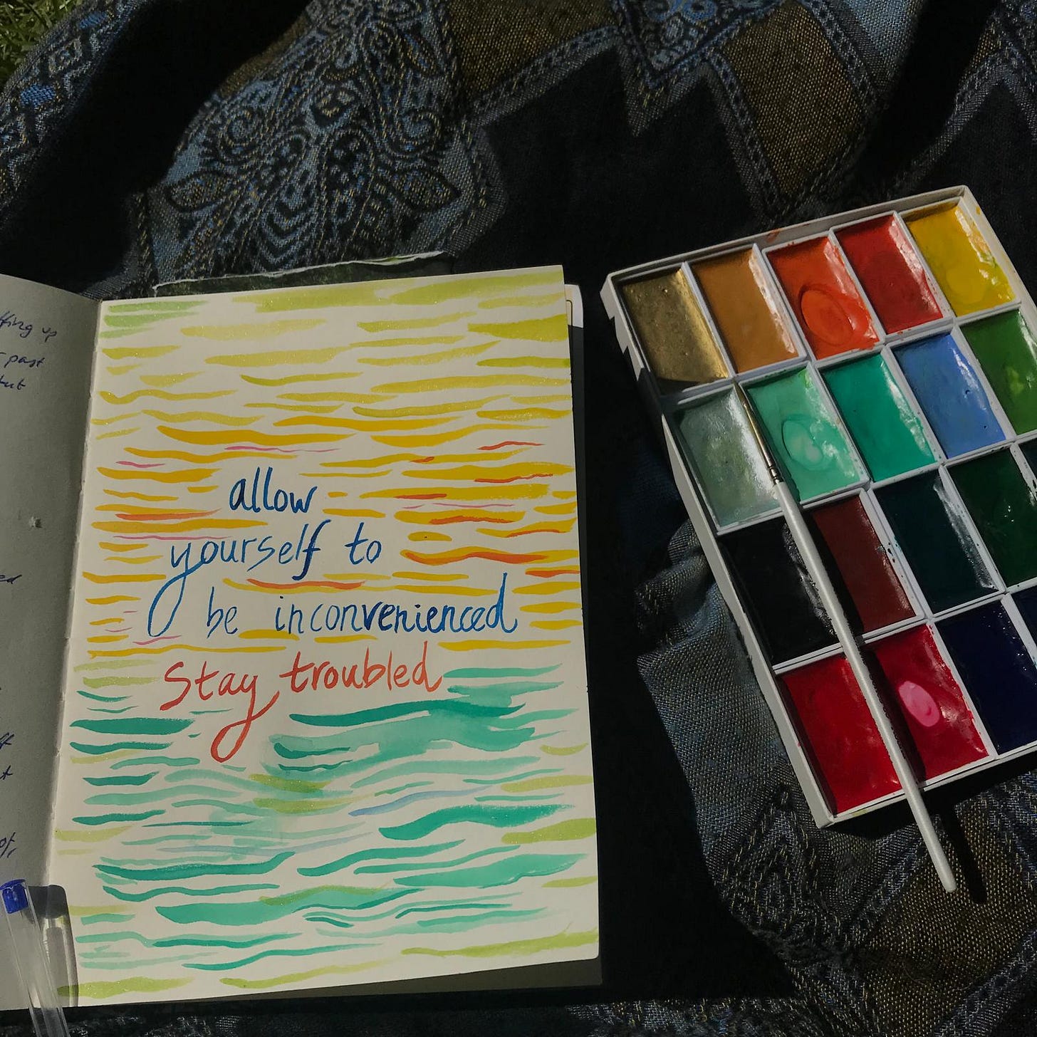 painted page that reads "allow yourself to be inconvenienced, stay troubled' which yellow waves as the background and a box of watercolour paints to the side, all on the background of a blue scarf