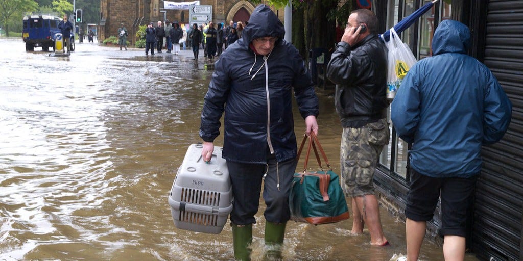 Man in waterproof coat and wellingtons walks through flooded street carrying a pet carrier and a holdall. Man in waterproof coat and wellingtons walks through flooded street carrying a pet carrier and a holdall.