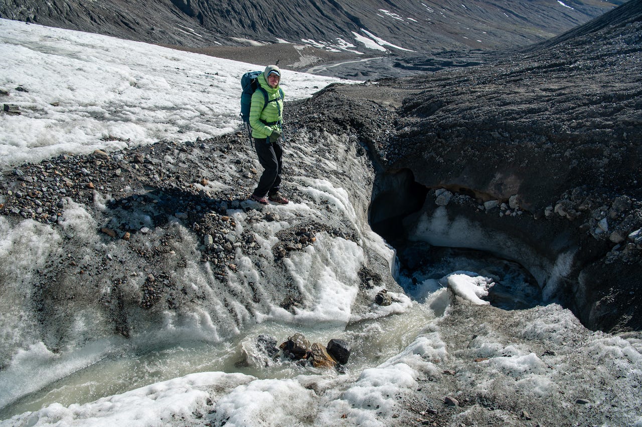 A hiker in a green puffy jacket and dark pants stands on the dirty, debris-strewn surface of a glacier, hands in pockets, beside a gap where meltwater has cut down into the ice and pools below. To the right, a dark hollow shows where the stream runs beneath the ice; a rubble-covered slope and remnant snow fill the valley behind under bright sun.