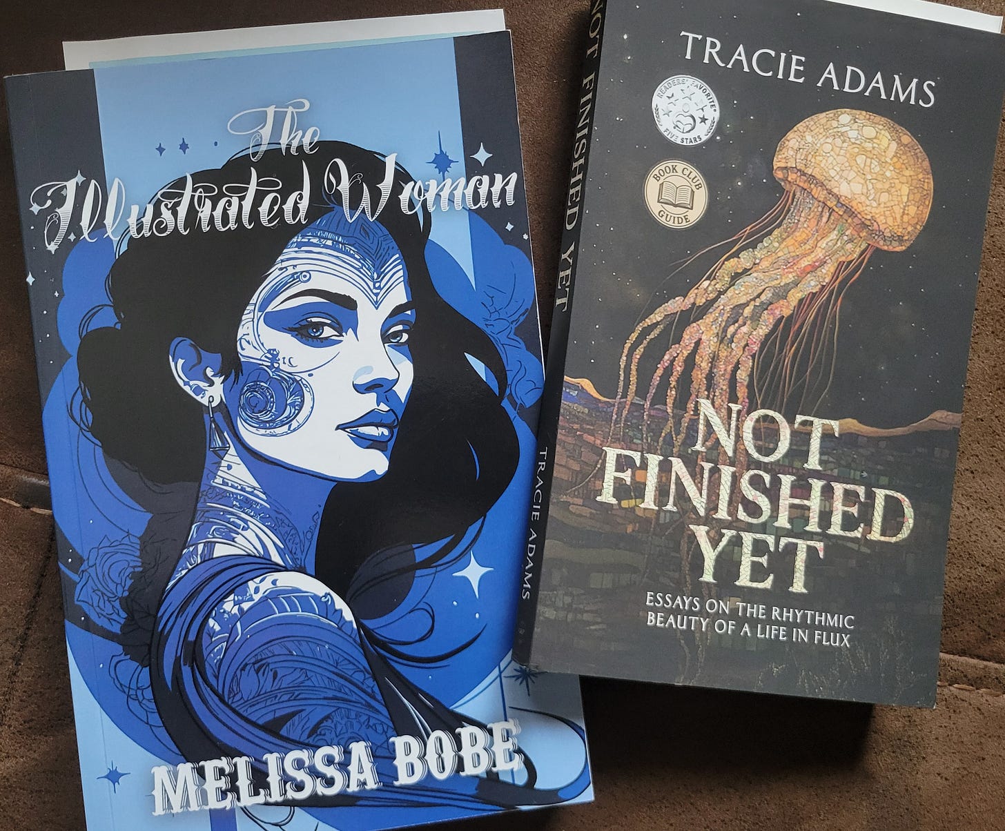 “The Illustrated Woman” by Melissa Bobe, and just delivered today “Not Finished Yet” by Tracie Adams. on the right, cover of jellyfish and on the right blue cover for Illustrated woman with woman with black hair and tattoos swirling on her face and neck