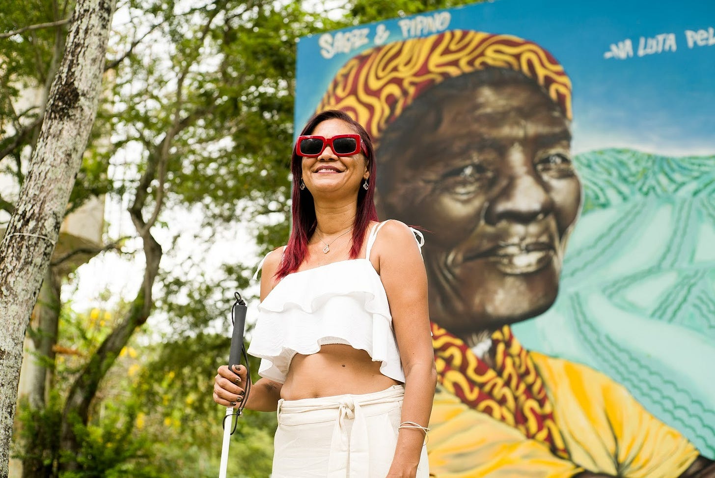 Cristiane aparece em um ambiente externo, durante o dia, sorrindo e de frente para a câmera. Ao fundo, há um grande mural colorido que retrata o rosto de uma mulher negra idosa com turbante, cercado por cores vibrantes e vegetação.