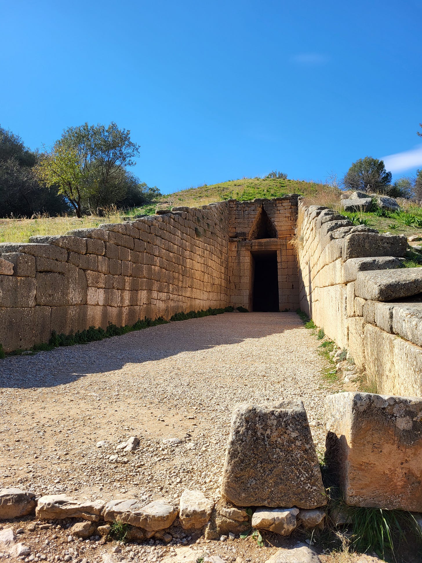 A stone brick structure built into a green hill in front of a blue sky. The entrance of the tomb is a a large empty rectangle with a triangle above it.