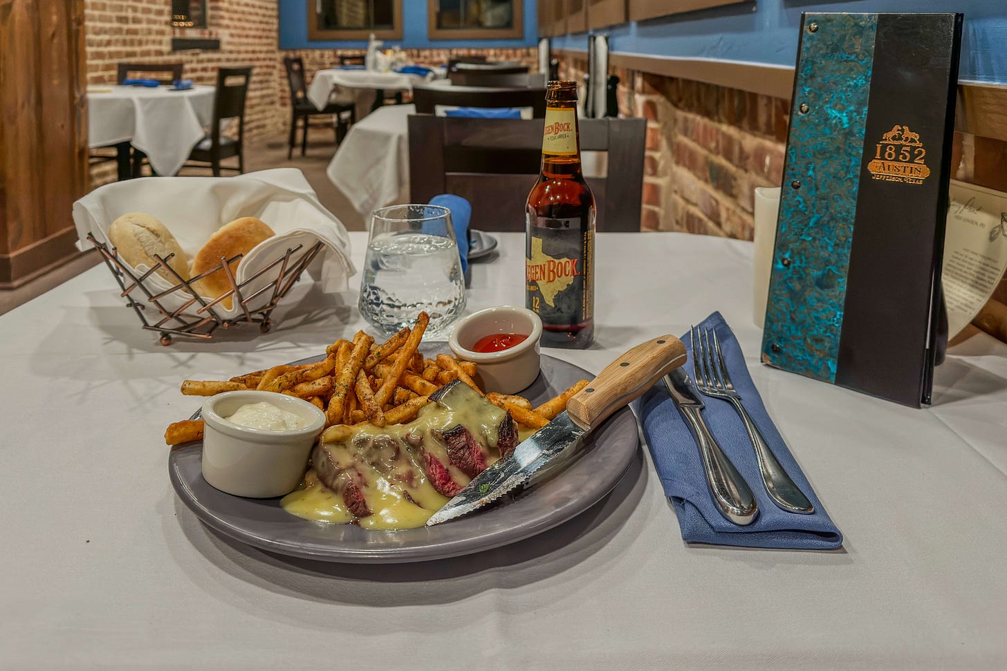Gedeckter Tisch im Restaurant «1852 on Austin» in Jefferson, Texas: Auf dem Teller liegt ein aufgeschnittenes Filet mit Sauce, dazu Pommes frites und Dips; daneben stehen Brot im Korb, ein Glas Wasser und eine Flasche Bier, im Hintergrund das rustikale Restaurantinterieur mit Backsteinwänden.