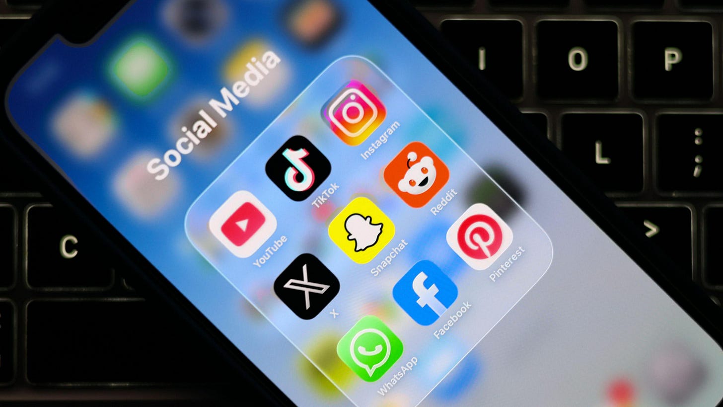 A mobile phone is held over a computer keyboard. It has a screen showing social media icons, including the black and white TikTok logo, green Whats App icon and yellow and black logo.