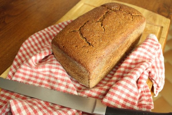 Amber loaf of ancient grain bread on a red plaid towel, on cutting board, with knife in foreground. 