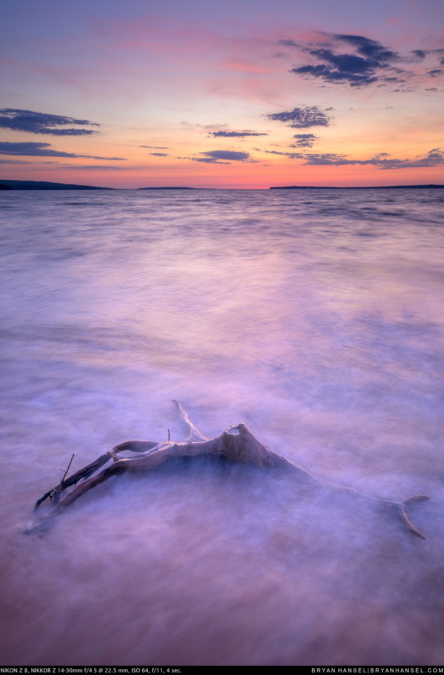 A weathered driftwood root emerges from silky smooth waves on Lake Superior at sunset, with pink and orange light glowing on the horizon and purple-tinted clouds above. Long exposure, 4 seconds.