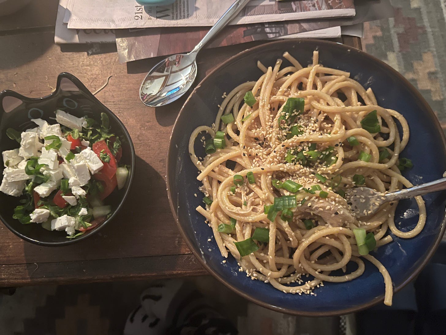 A small cat bowl filled with salad and a large blue bowl filled with pasta and a fork atop a wooden tabletop with a silver spoon nearby. A small cat bowl filled with salad and a large blue bowl filled with pasta and a fork atop a wooden tabletop with a silver spoon nearby.