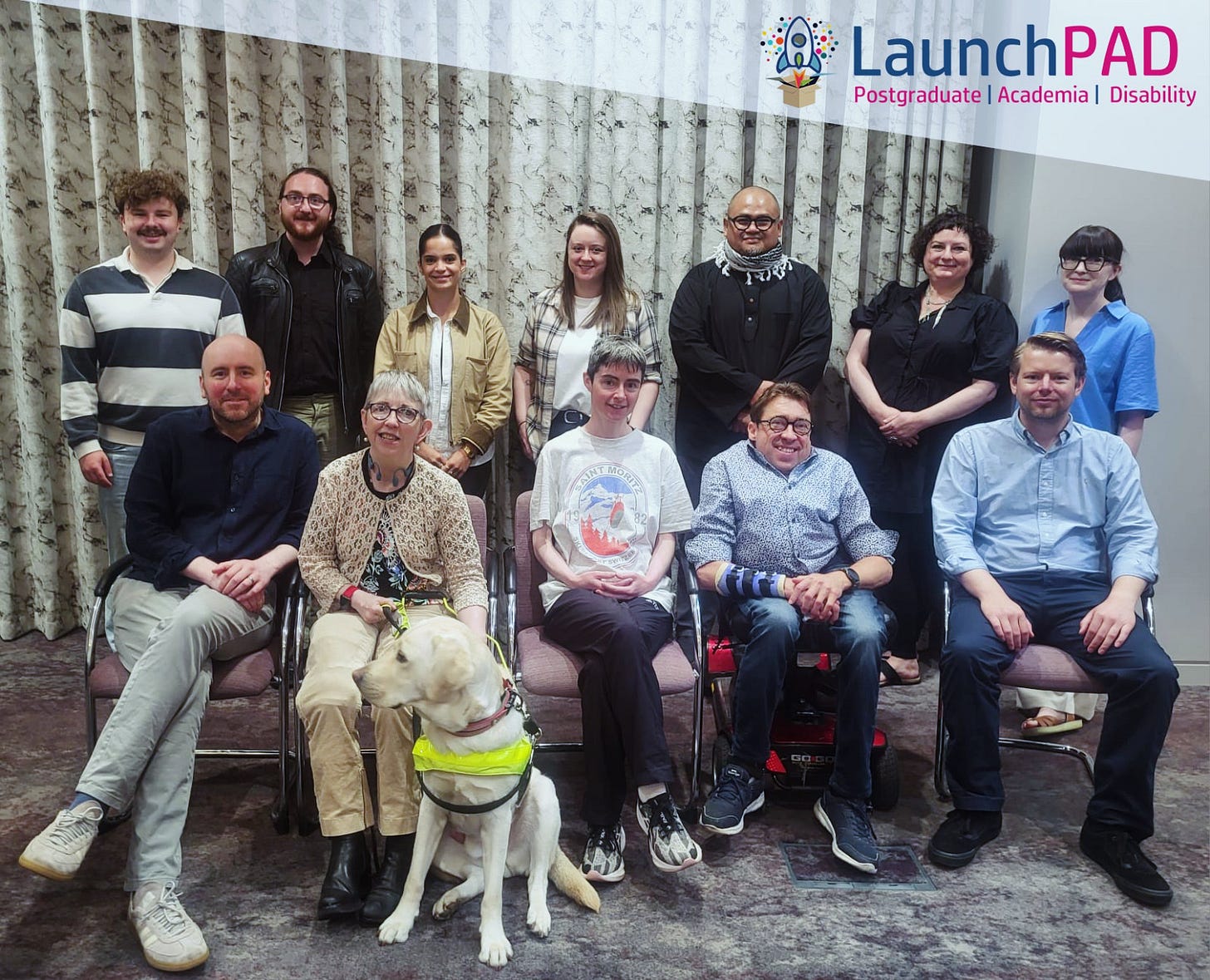 Group photograph of approximately eleven LaunchPAD participants at an indoor event space, posed in two rows in front of patterned curtains. The LaunchPAD logo appears in the top right corner with the tagline “Postgraduate | Academia | Disability”. The front row includes seated participants, among them two wheelchair users and a guide dog wearing a yellow harness. The back row consists of standing participants in casual and semi-formal attire. One participant in black traditional attire stands near the centre-right. The group appears relaxed and smiling, reflecting an inclusive postgraduate disability community gathering.