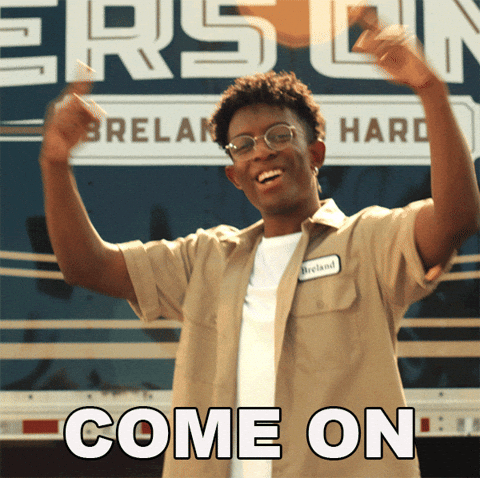 This image is a GIF from the music video for the song "Beers On Me" by Dierks Bentley featuring Breland and Hardy. A Black man smiles warmly and gives an enthusiastic gesture with his arms raised and circling his pointer fingers while nodding approvingly.