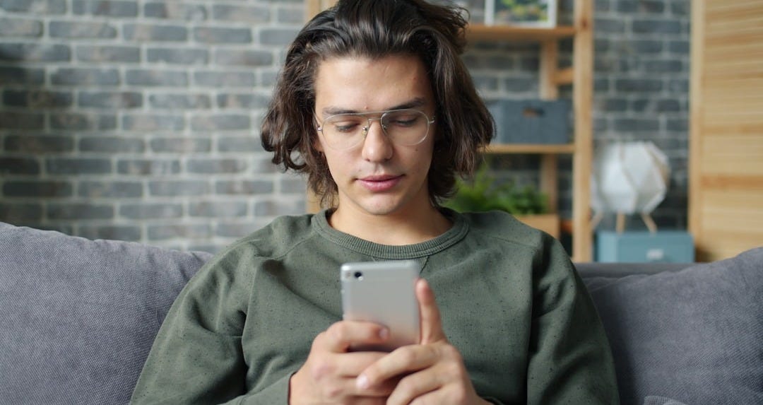 Young man with glasses using a smartphone on couch.