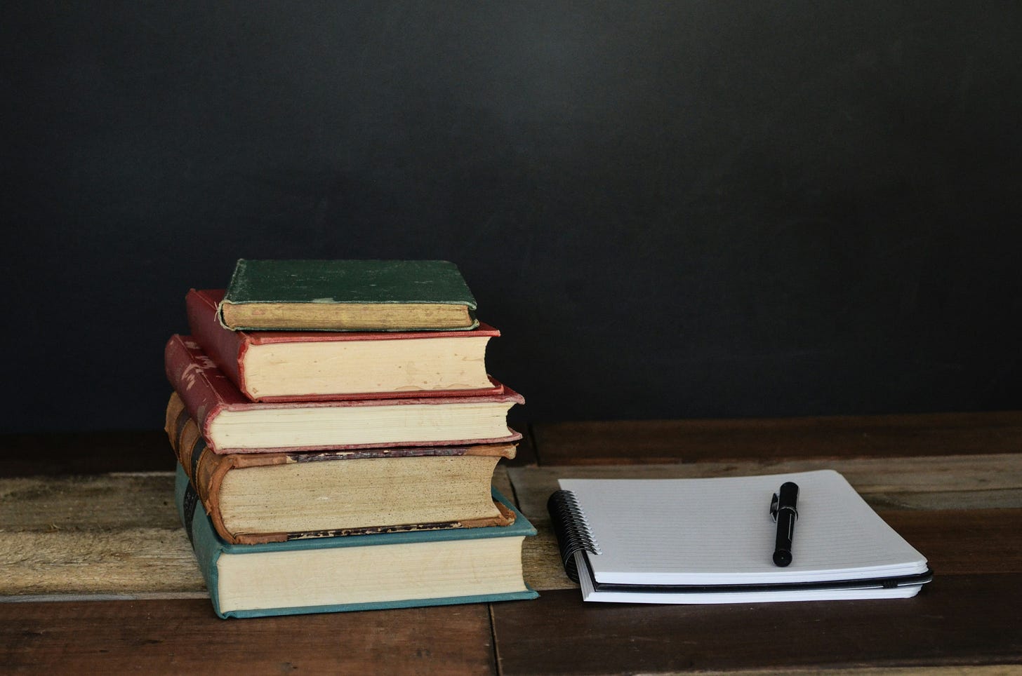 A stack of old, weathered books sits on a wooden table beside an open spiral notebook with a black pen resting on top, evoking quiet study and thoughtful reflection. A stack of old, weathered books sits on a wooden table beside an open spiral notebook with a black pen resting on top, evoking quiet study and thoughtful reflection.