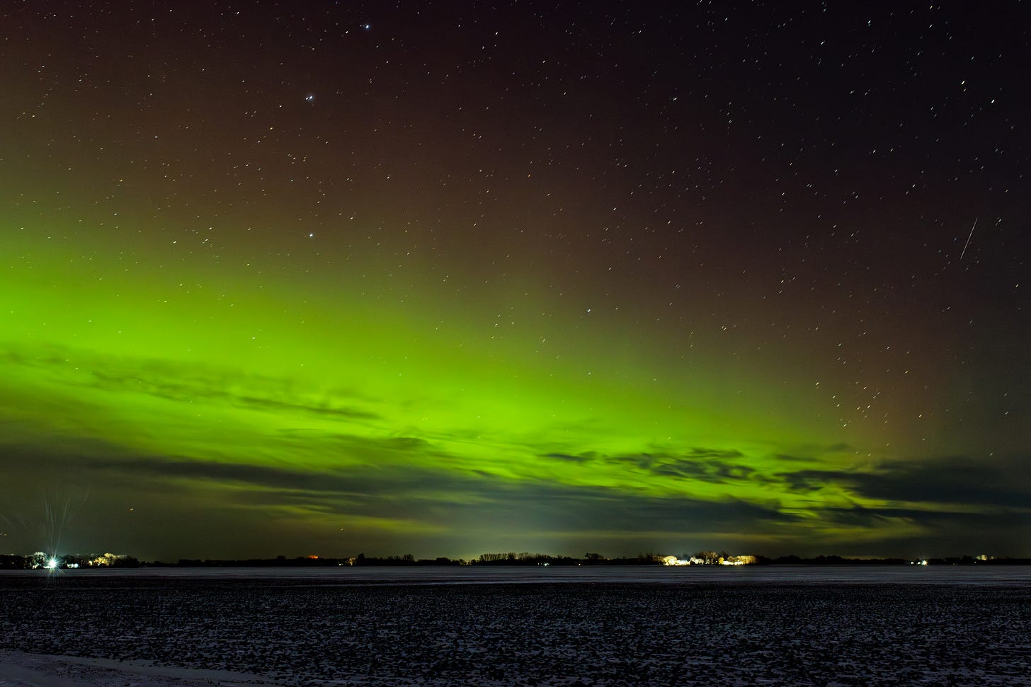 a farm field with clouds above it and the bright green, red-topped glow of the aurora borealis with clouds at the bottom. there is a bright  meteor on the right.
