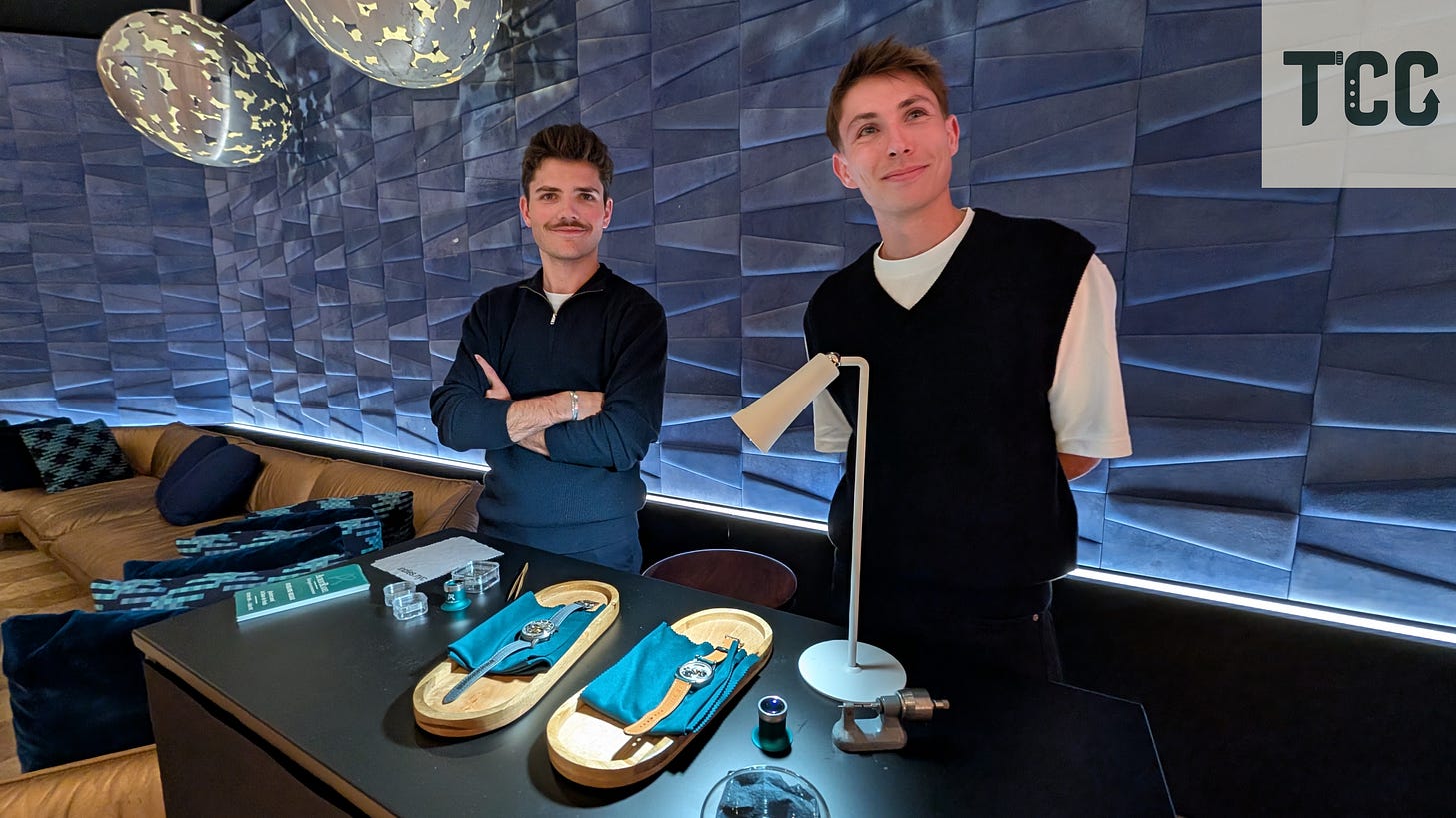 Two young men standing behind a watch display table at The Indies. The table has two watches on padded trays, magnifying loupes, and a compact desk lamp. The background features a textured blue wall with modern pendant lights.