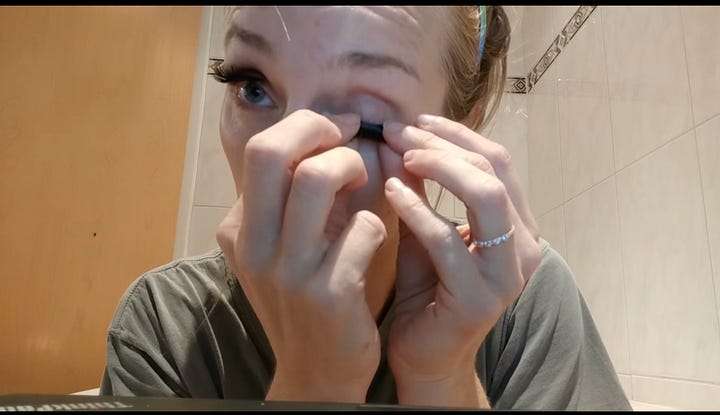 Primo piano su una persona che si trucca o applica ciglia finte sul suo occhio destro. Usa le dita e uno strumento nero per tenere ferma la palpebra e posizionare l'oggetto scuro vicino alla linea delle ciglia. La persona indossa una maglietta grigia, ha lunghe ciglia scure e un anello sottile con piccole pietre al dito medio della mano destra.