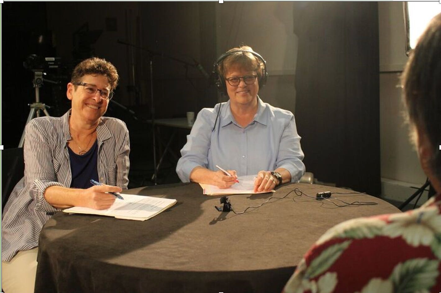 In a studio recording setting, two people sit at a round table taking notes. The person seated on the left writes on a notepad, while the person on the right wears over‑ear headphones and writes on paper. Small microphones rest on the table, with studio lighting and a camera tripod visible in the background. A third person is partially visible in the foreground, facing the table.