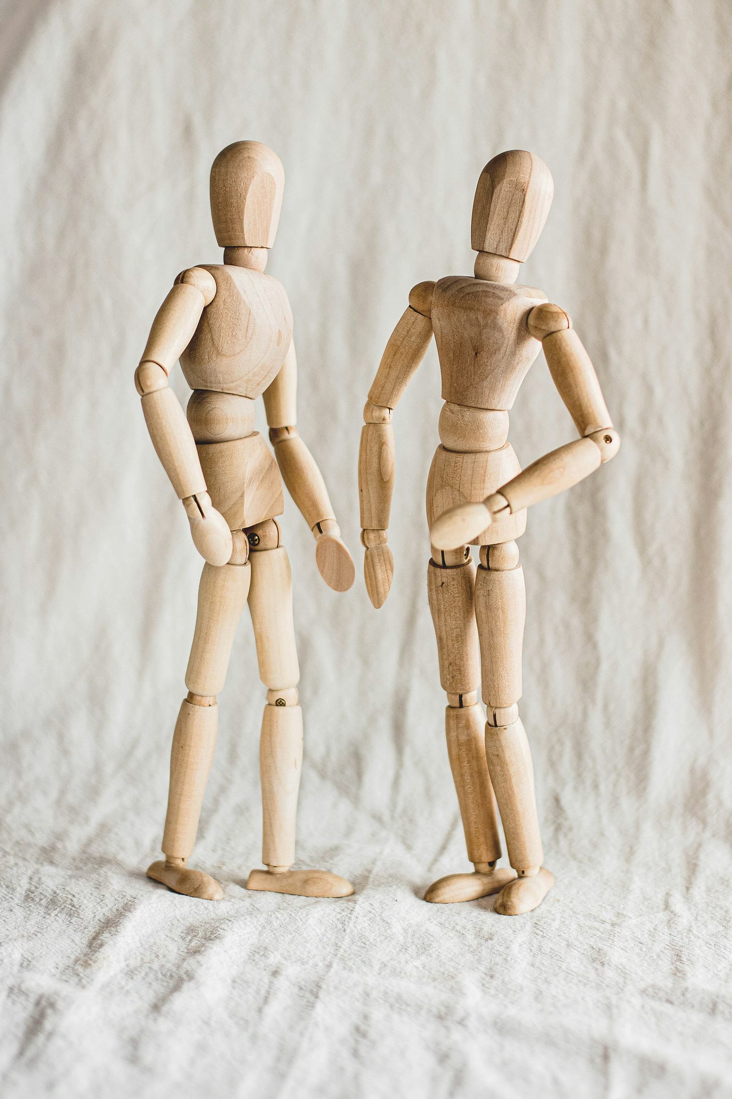 Two light colored wooden human figures, with jointed limbs but no faces or other detail, standing in front of a beige tarp. Two light colored wooden human figures, with jointed limbs but no faces or other detail, standing in front of a beige tarp.