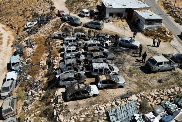 An overhead view of a few people standing near two buildings and a cluster of scorched vehicles.