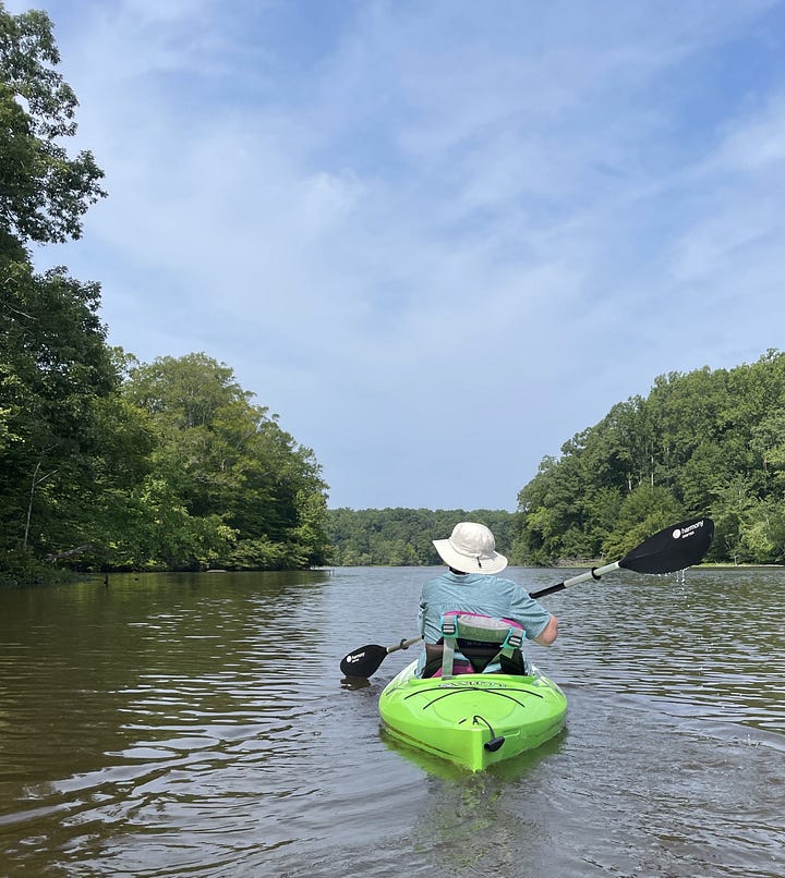 on left: person in green shirt and white hat, paddling a green kayak, trees visible on both sides, blue sky. On right: person in blue and black shirt and black shorts standing at end of kayak, feet in the water on either side of kayak, holding a cane and staff behind back. Two stools are beside kayak on ground: a plastic step stool and a taller garden stool.
