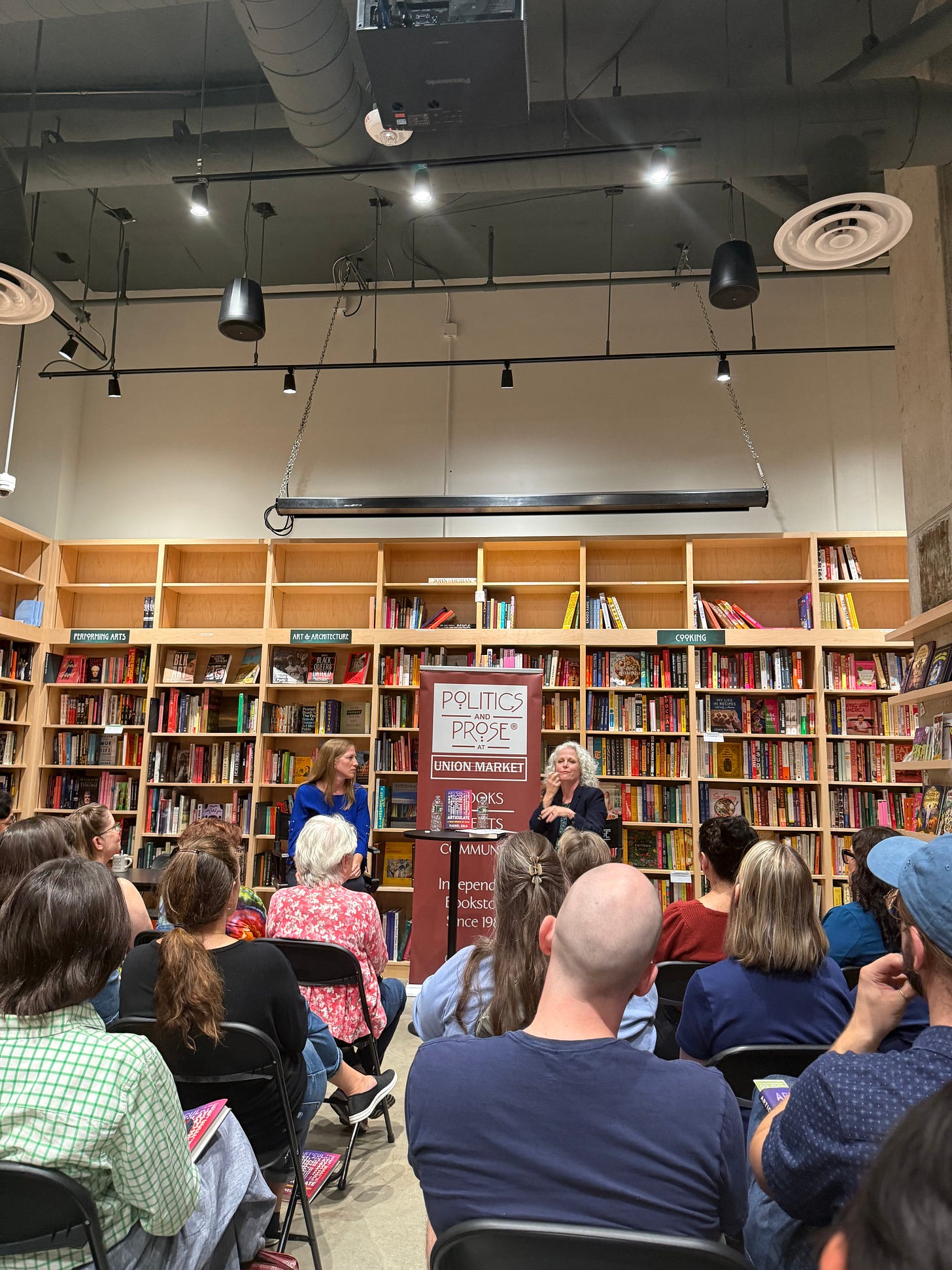 Photo taken from the audience in Politics and Prose at Union Market. I'm sitting at the front of the bookstore with Kristen Harmon, who's in the middle of signing something. Bookshelves are in the background, seated audience members in the foreground.