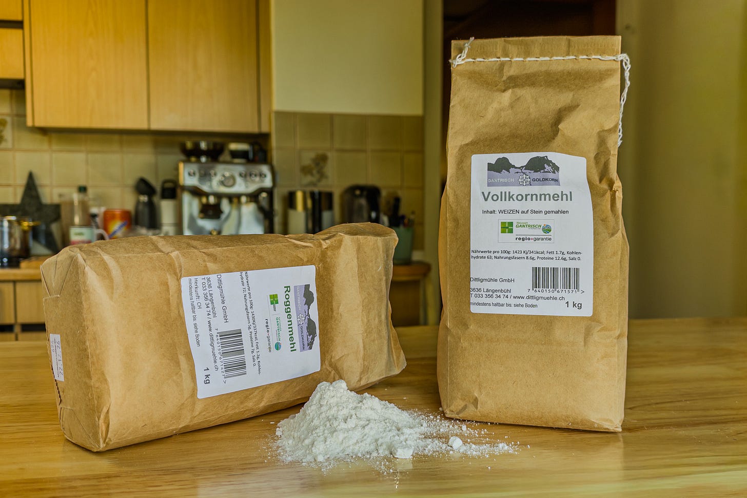 Zwei braune Papierbeutel mit je einem Kilogramm Mehl stehen auf einem Holztisch in einer Küche. Links liegt eine Packung Roggenmehl, rechts steht eine Packung Weizenvollkornmehl der Dittligmühle aus dem Gantrischgebiet. Dazwischen liegt ein kleiner Haufen helles Mehl. Im Hintergrund sind Küchenschränke und eine Kaffeemaschine zu sehen.