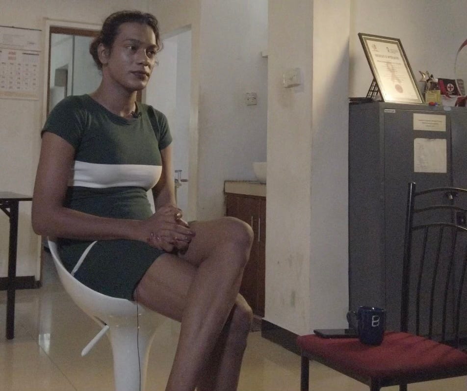 A slender person with dark, pulled back hair, sits with legs crossed on a white chair