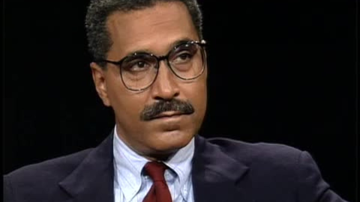 Shelby Steele wearing glasses, a mustache, a dark suit jacket, a light blue dress shirt, and a red tie, sitting against a black background.