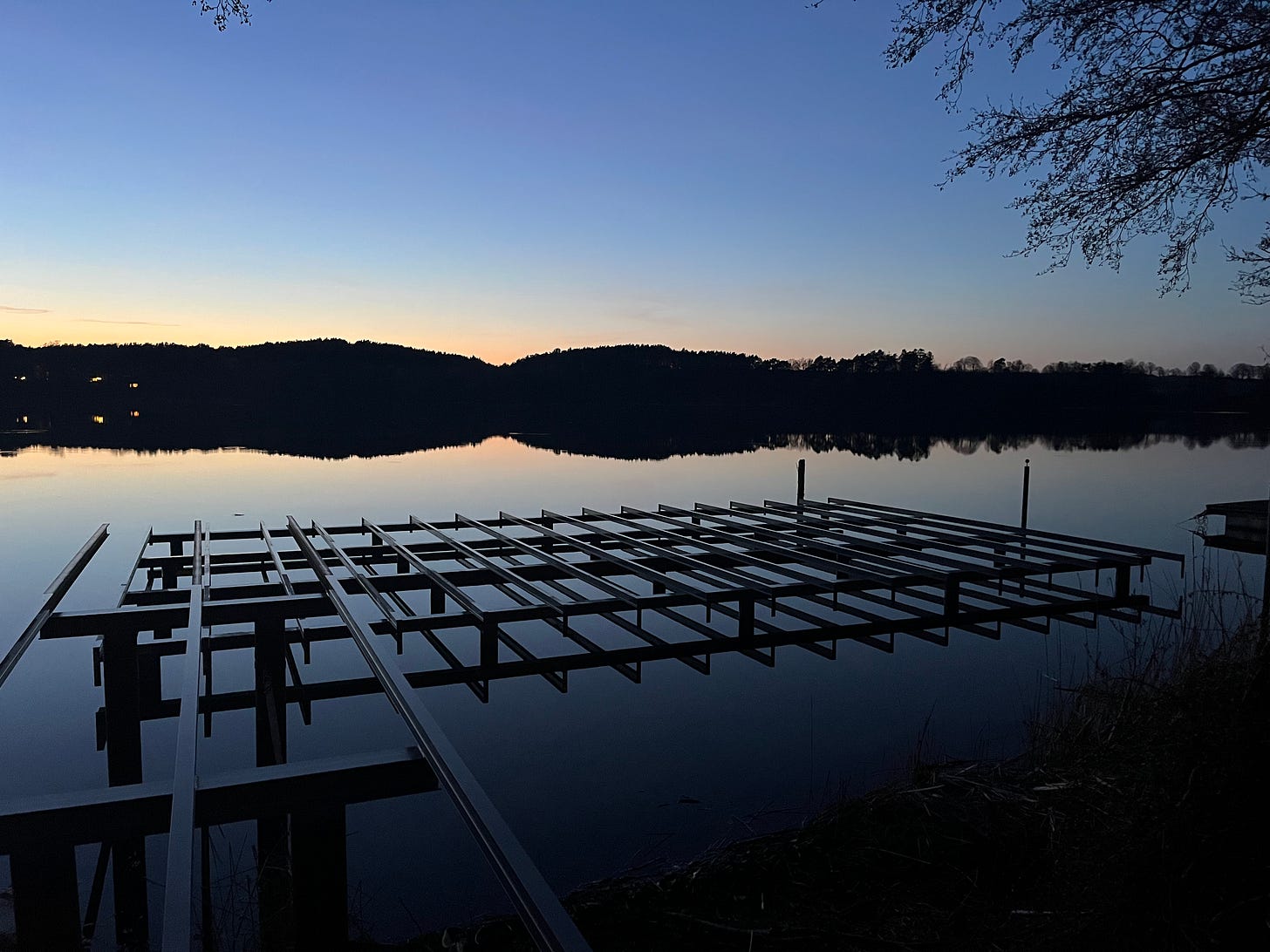 Ein See im letzten Abendlicht, über der Oberfläche ein Gerüst aus Metallstreben für einen neuen Steg, das sich im ruhigen Wasser spiegelt