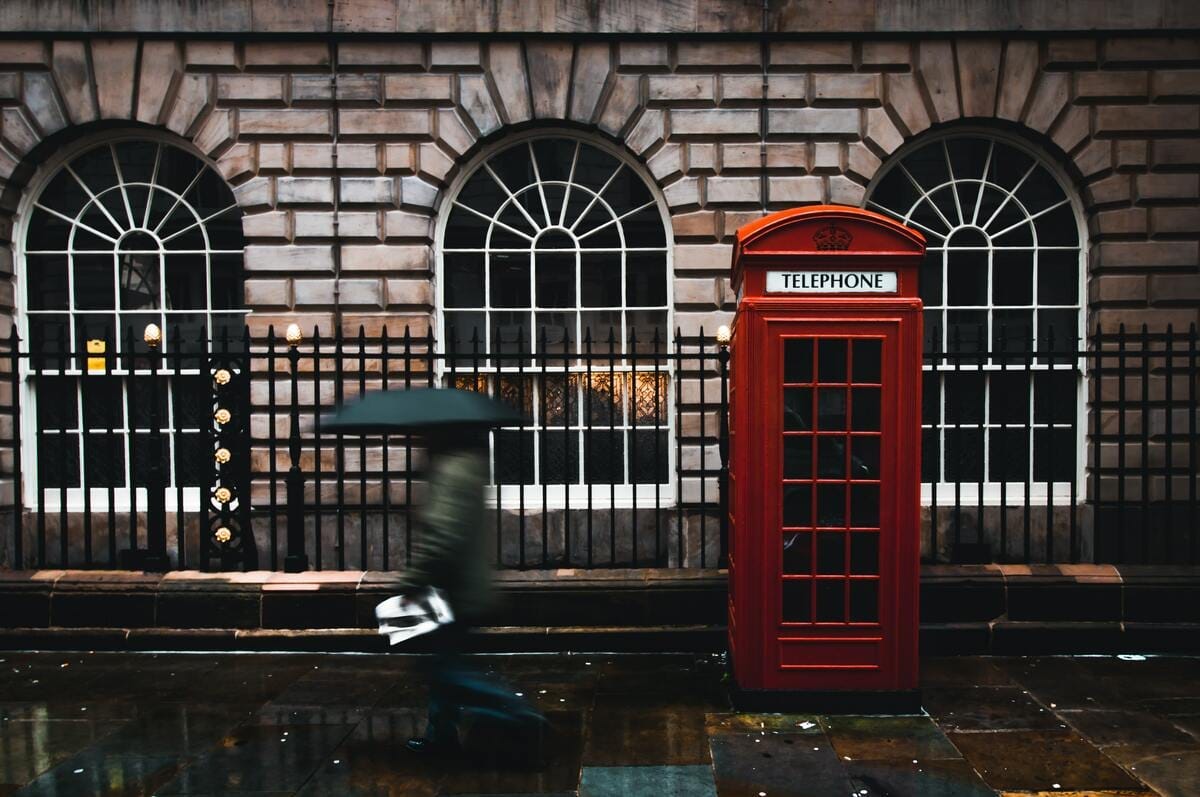 Exterior image of a building in London and the red telephone booth outside it with a pedestrian crossing, carrying an umbrella