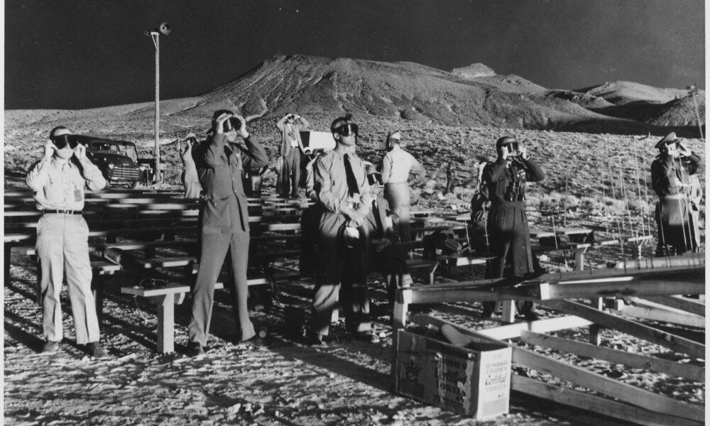 black and white photo of people observing a nuclear test explosion in the Nevada desert in 1955; they all hold goggles to their faces to protect themselves from the flash of the blast black and white photo of people observing a nuclear test explosion in the Nevada desert in 1955; they all hold goggles to their faces to protect themselves from the flash of the blast