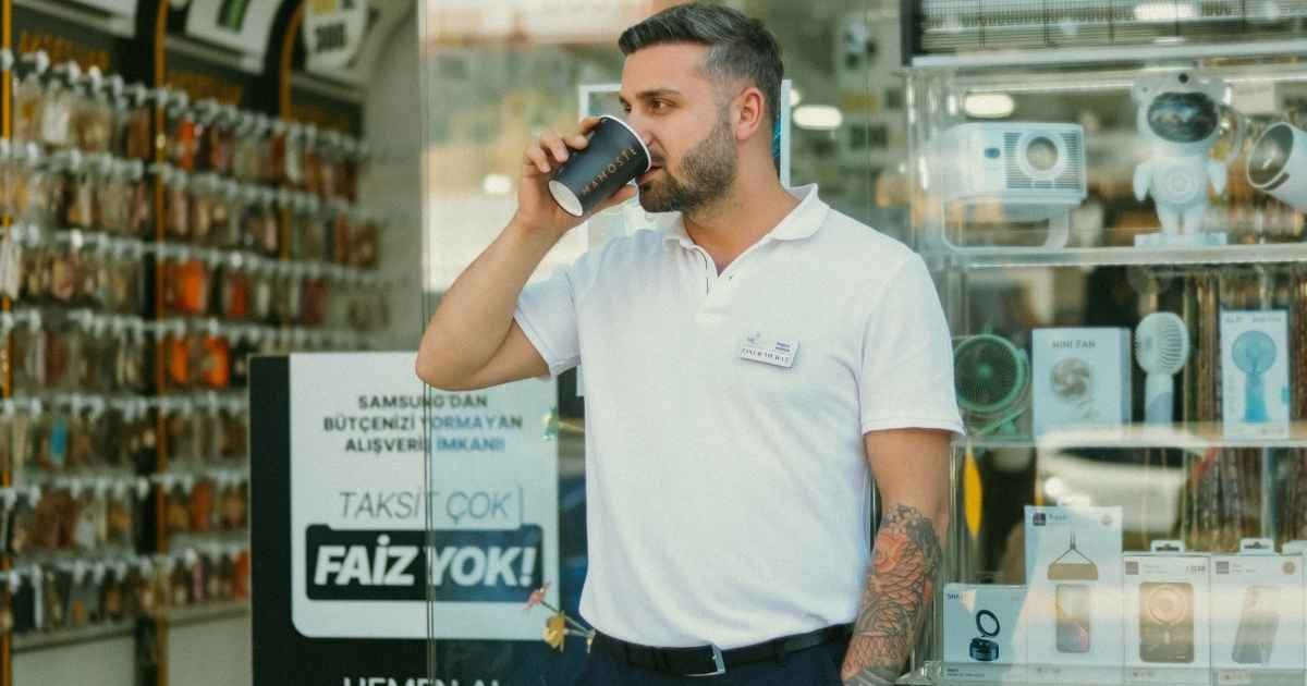 Young man sips coffee while standing in front of an electronics shop, surrounded by gadgets.