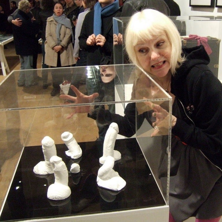 Cynthia Allbritton, a woman with white shoulder-length hair, poses happily next to a glass case containing white plaster phalluses
