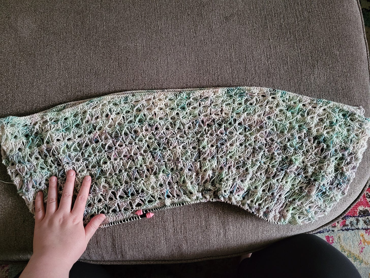 A panel of knitted broomstick lace in a watercolor-style variegated yarn in greens, blues, purples, and white. The panel is spread out on a gray ottoman and a white woman's hand is on top of it. A panel of knitted broomstick lace in a watercolor-style variegated yarn in greens, blues, purples, and white. The panel is spread out on a gray ottoman and a white woman's hand is on top of it.