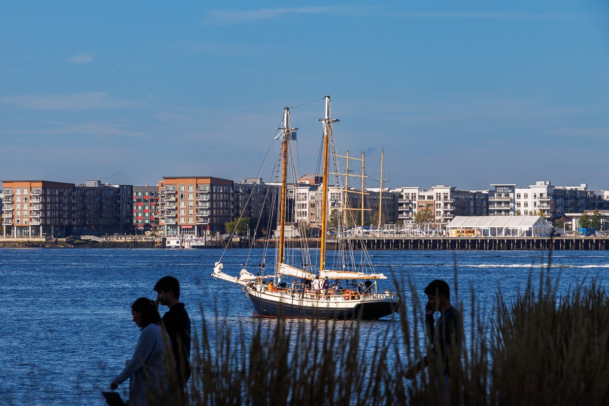 Two-masted schooner cruising in Boston Harbor with apartment buildings behind and silhouetted pedestrians in front Two-masted schooner cruising in Boston Harbor with apartment buildings behind and silhouetted pedestrians in front