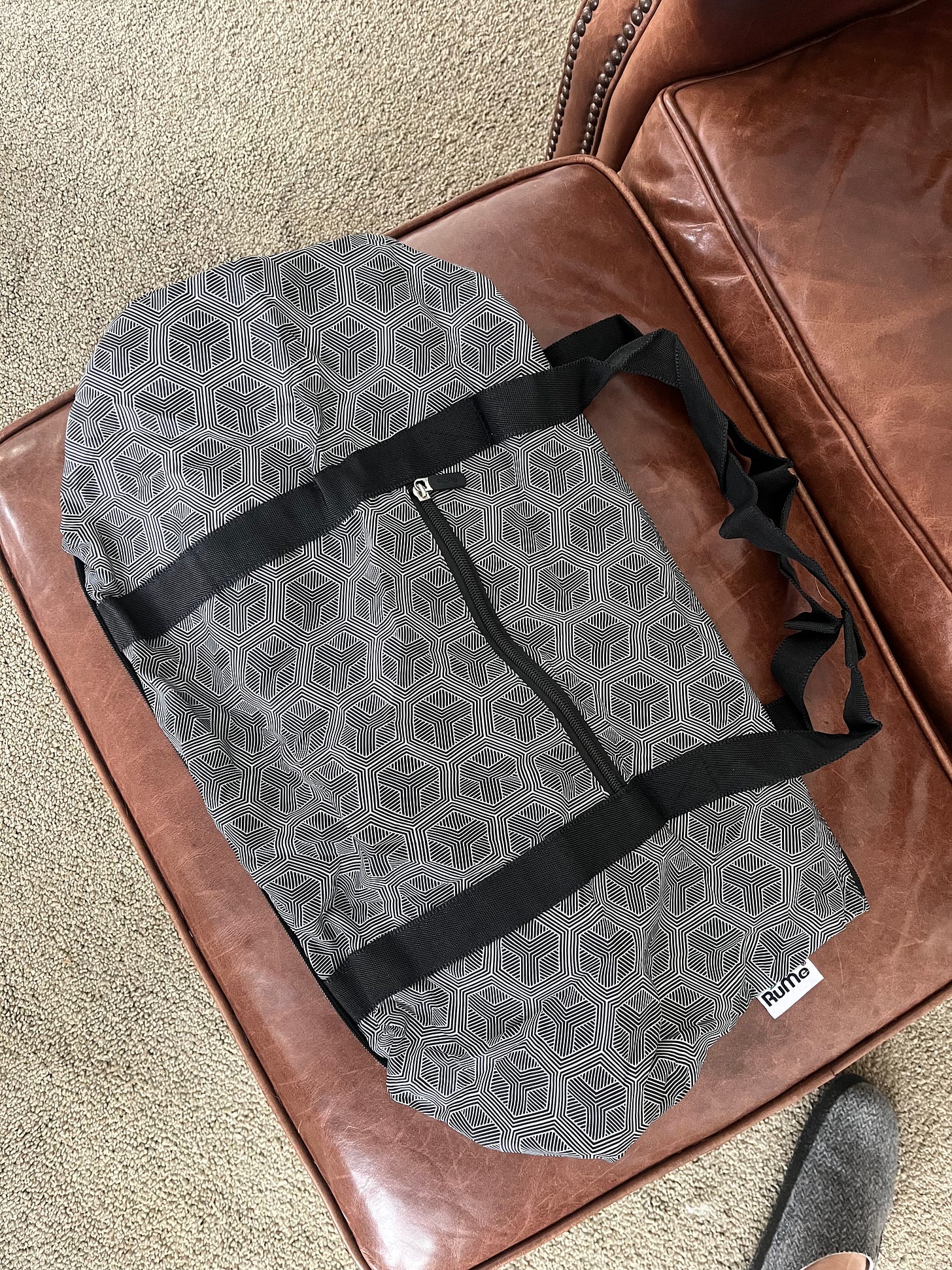 a black and white patterned shoulder bag sits on top of a leather ottoman. Also pictured is a small portion of a similarly white and black patterned slipper. a black and white patterned shoulder bag sits on top of a leather ottoman. Also pictured is a small portion of a similarly white and black patterned slipper.